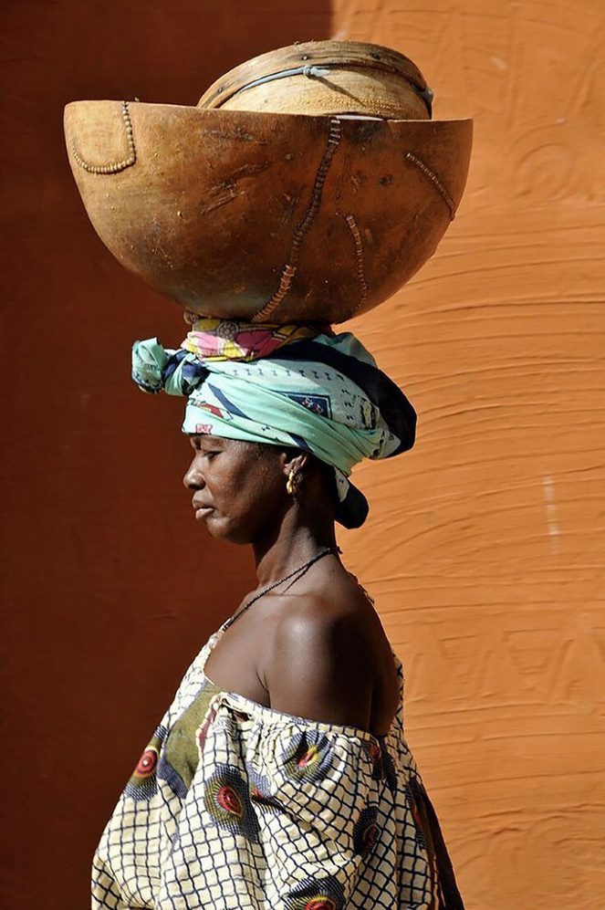 Femme portant une calebasse sur la tête devant un mur ocre